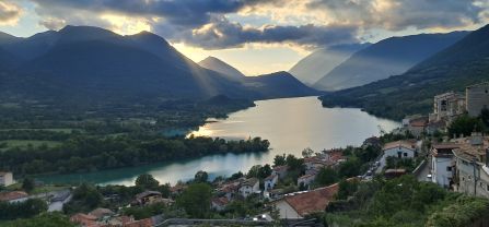A lake in front of beautiful mountains. The sun shines through the clouds and a human settlement is seen in the  in the foreground.