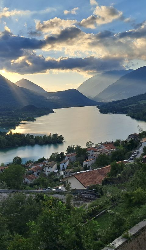 A lake in front of beautiful mountains. The sun shines through the clouds and a human settlement is seen in the  in the foreground.
