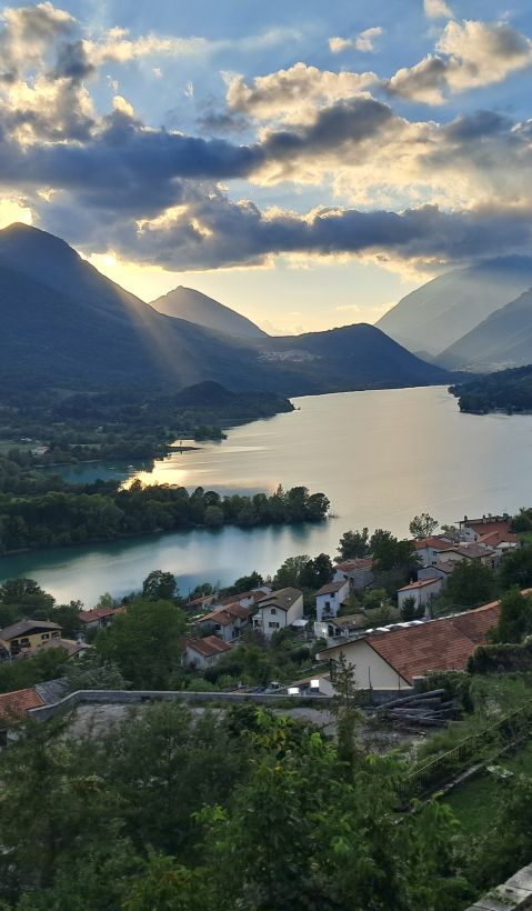 A lake in front of beautiful mountains. The sun shines through the clouds and a human settlement is seen in the  in the foreground.