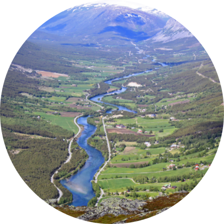 An aerial picture of Gudbrandsdalen valley. A blue river if flowing though the green landscape. Snowy mountains are seen in the background and small clusters of human settlements are scattered at the bottom of the valley. 